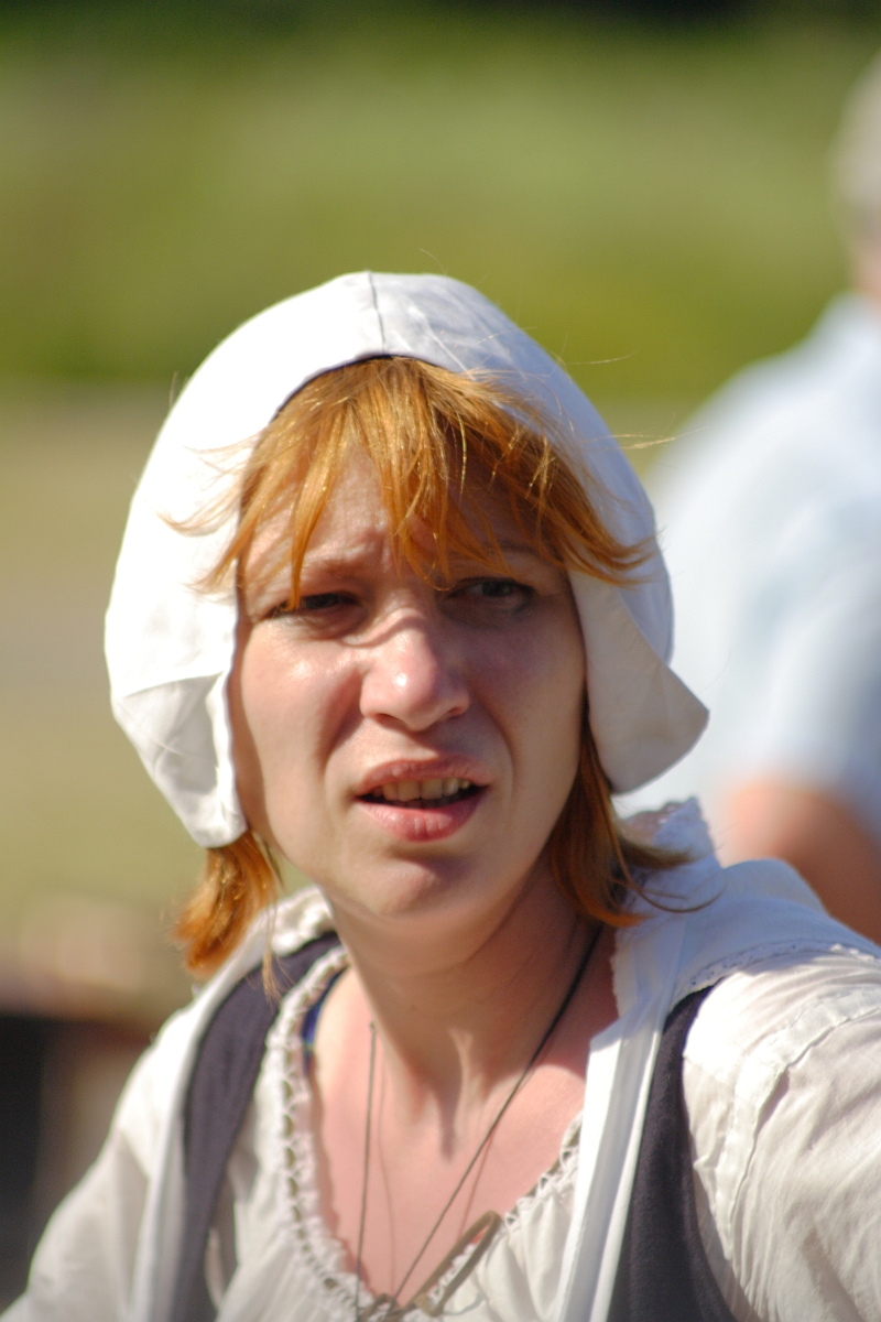 A woman with red hair wears a white bonnet and a white blouse with a black vest. Outdoors in a sunny environment with blurred greenery in the background, her facial expression suggests concern or confusion, reminiscent of an enigmatic figure from Dunamunde 2010.