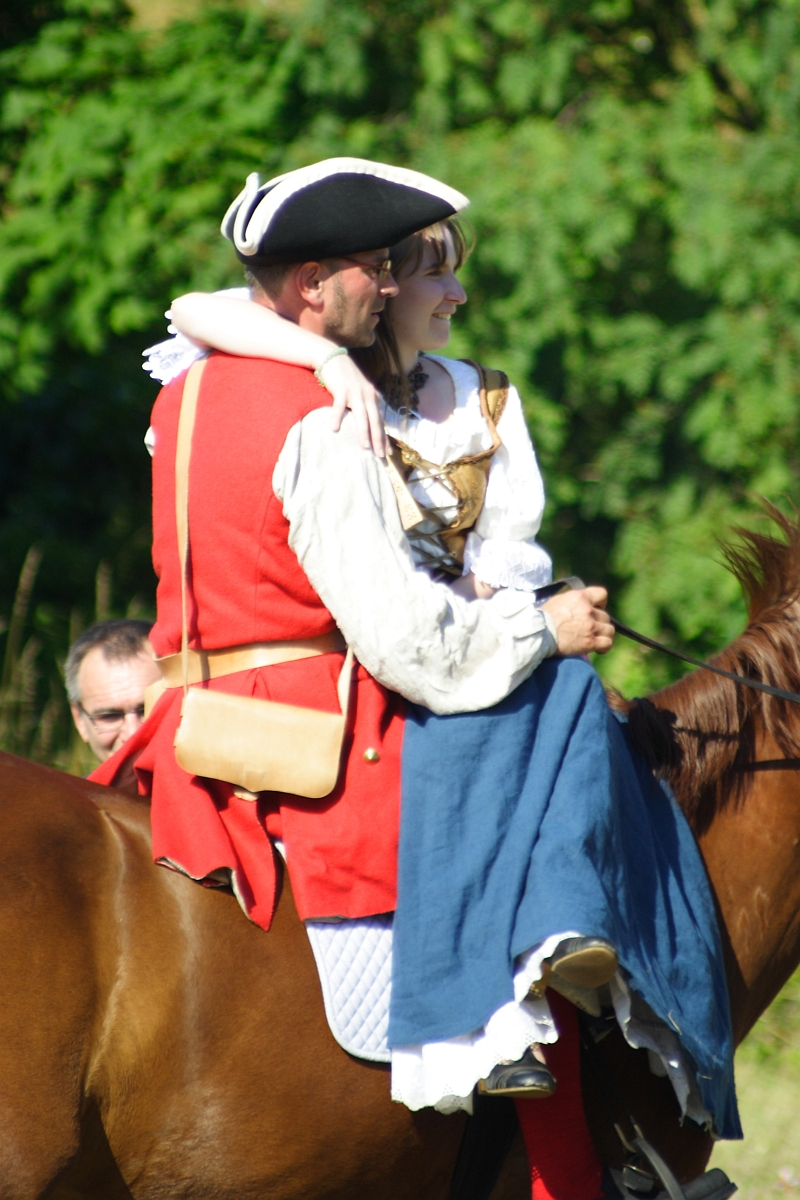 Two people dressed in historical attire ride a brown horse at the Dunamunde 2010 reenactment. The person in front wears a red coat, white shirt, and black tricorne hat, while the person behind has a white blouse and blue dress. They embrace warmly. The backdrop is lush and green, suggesting an outdoor setting.