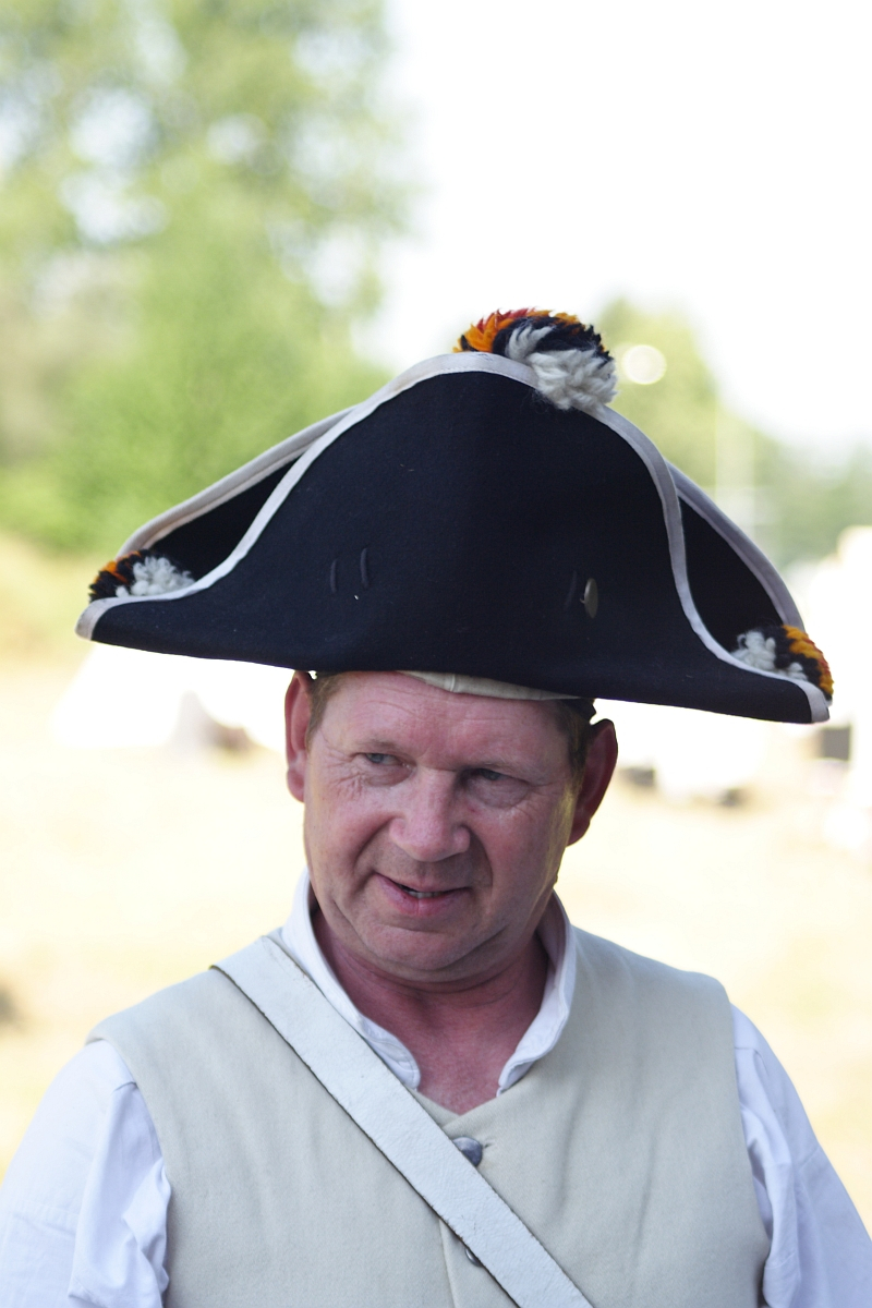 A middle-aged man with light hair is dressed in a historical costume, which includes a white vest over a white shirt. He wears a large, black tricorne hat adorned with small pom-poms in white, orange, and black. The background is blurred with greenery and light at Dunamunde 2010.