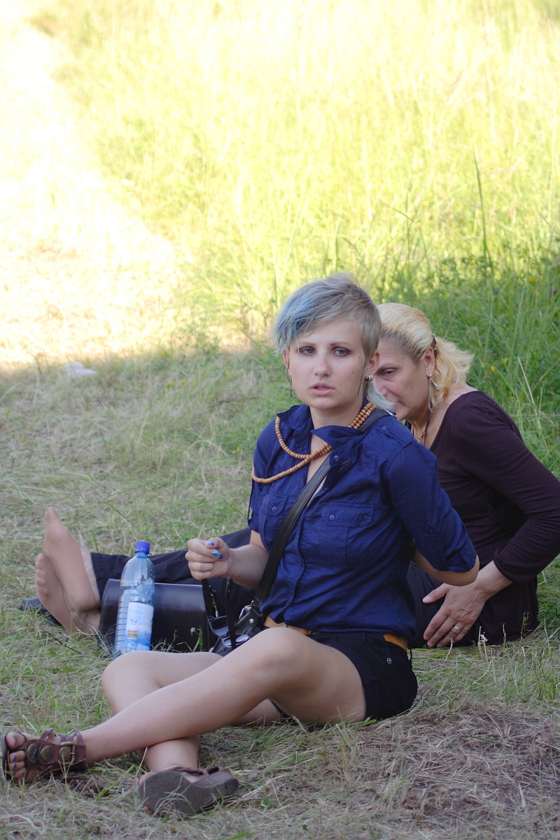 Two women are sitting on the grass, one in front of the other. The woman in front, resembling a 2010 fashion trend with her short, light blue hair, is wearing a blue shirt, black shorts, and sandals while holding a phone. Behind her, a blonde woman in a brown shirt and pants leans forward. A water bottle rests on the ground beside them.