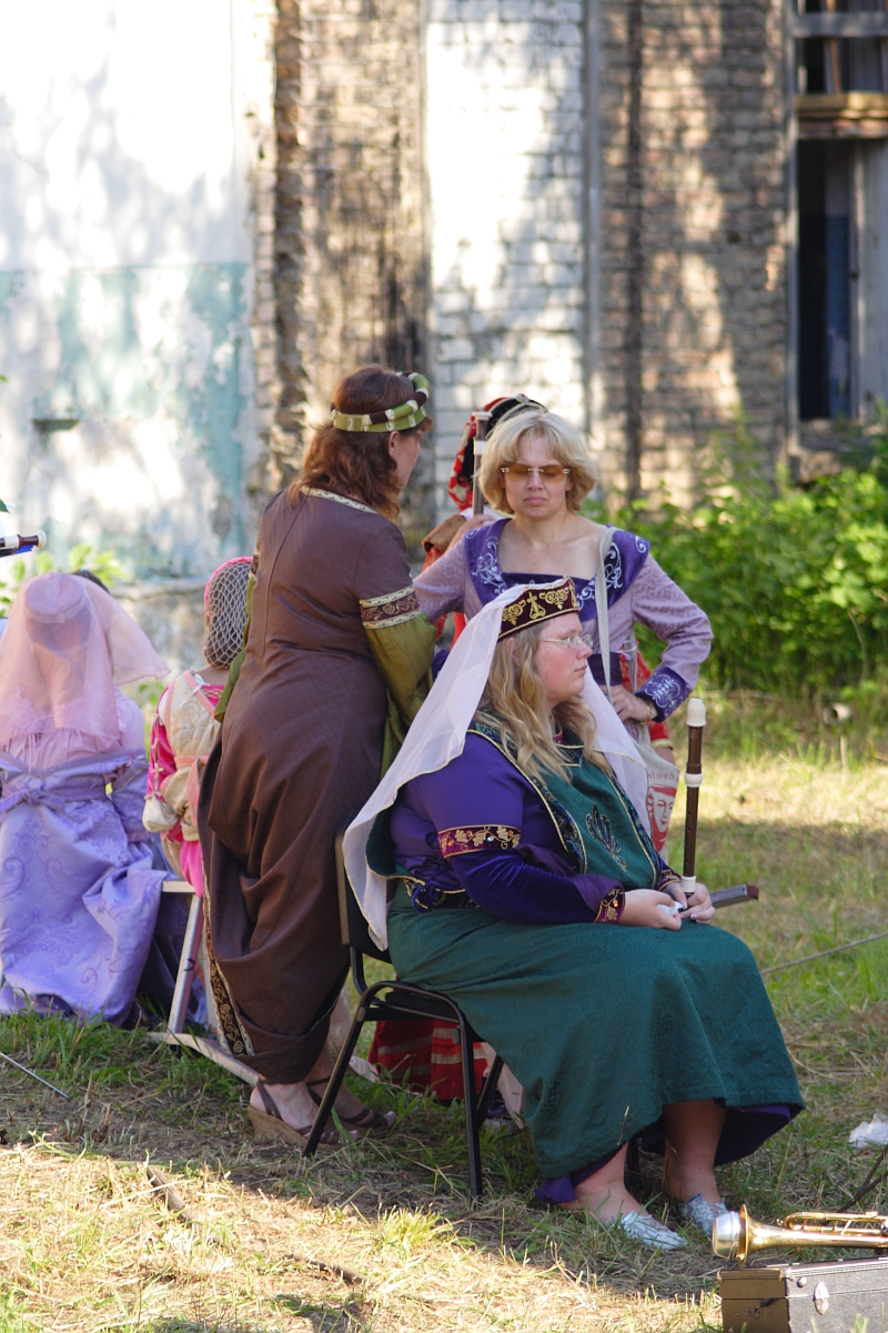 A group of people dressed in medieval attire, reminiscent of a 2010 Dunamunde festival, is depicted outdoors. One person sits on a chair in green and purple robes with a white veil, while another in a brown dress and flower crown stands nearby. A third person in purple and blue garments and wearing glasses also stands. Trees and a building are in the background.