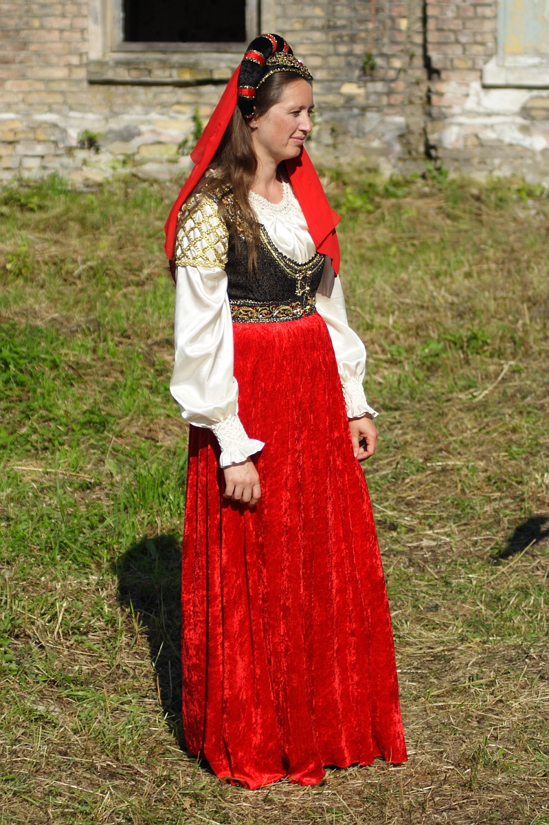 A woman stands on grass wearing traditional attire featuring a long red velvet skirt, a white blouse with puffed sleeves, and a black and gold embroidered vest. She also has a red headscarf with golden accents draped over her hair. The background shows a weathered brick wall and a window, reminiscent of Dunamunde 2010.