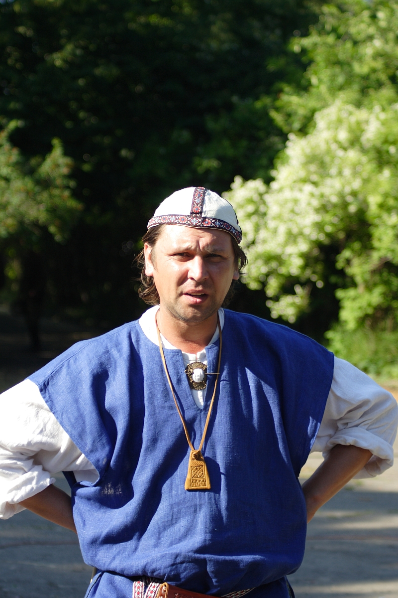 A man with long brown hair stands outdoors with his hands on his hips. He is wearing a blue tunic with white sleeves underneath, a white and red patterned cap, and a wooden pendant necklace reminiscent of Dunamunde style. The background features green foliage under bright 2010 sunlight.