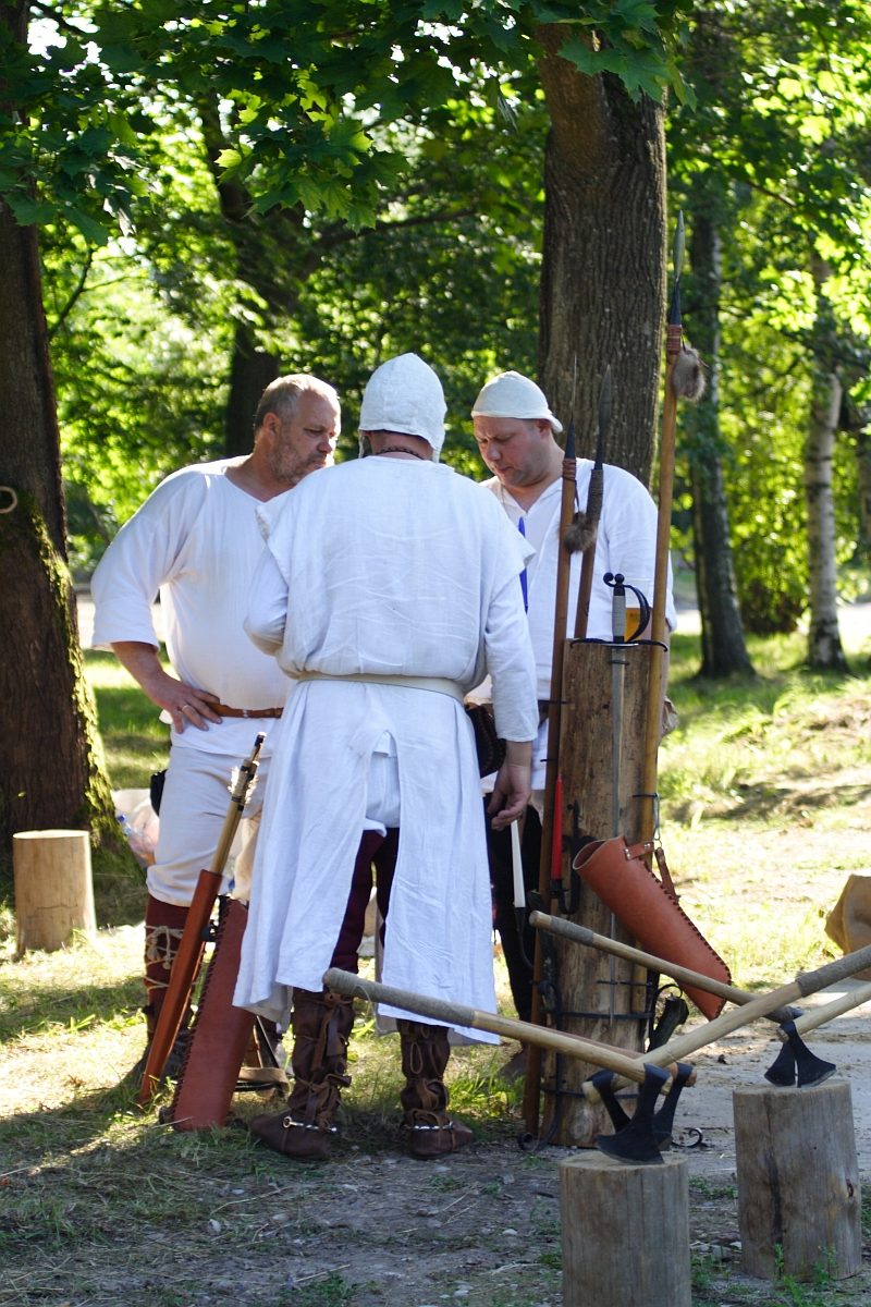 Three men dressed in medieval white tunics and caps stand in a wooded area at Dunamunde, engaged in conversation. Various medieval tools and weapons, including an axe and sword, are displayed on a wooden stand nearby. The scene is bright and sunny, with green trees providing a backdrop reminiscent of 2010.