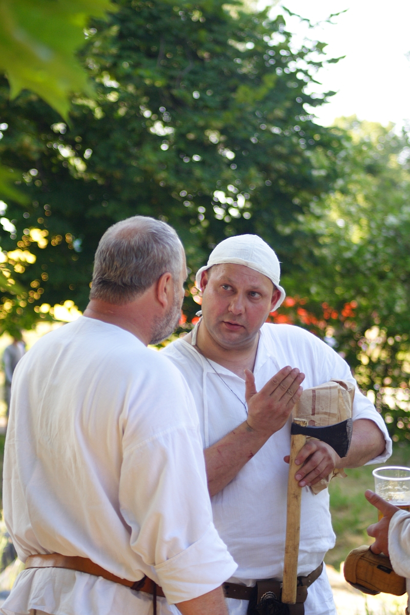 Two men in historical attire are conversing outdoors in Dunamunde. Both wear white tunics; the man on the right also wears a white head covering and holds an axe with a cloth on the blade. They stand in a green, leafy setting, seemingly engaged in discussion. A blurred person is in the background, hinting at 2010 reenactment activities.