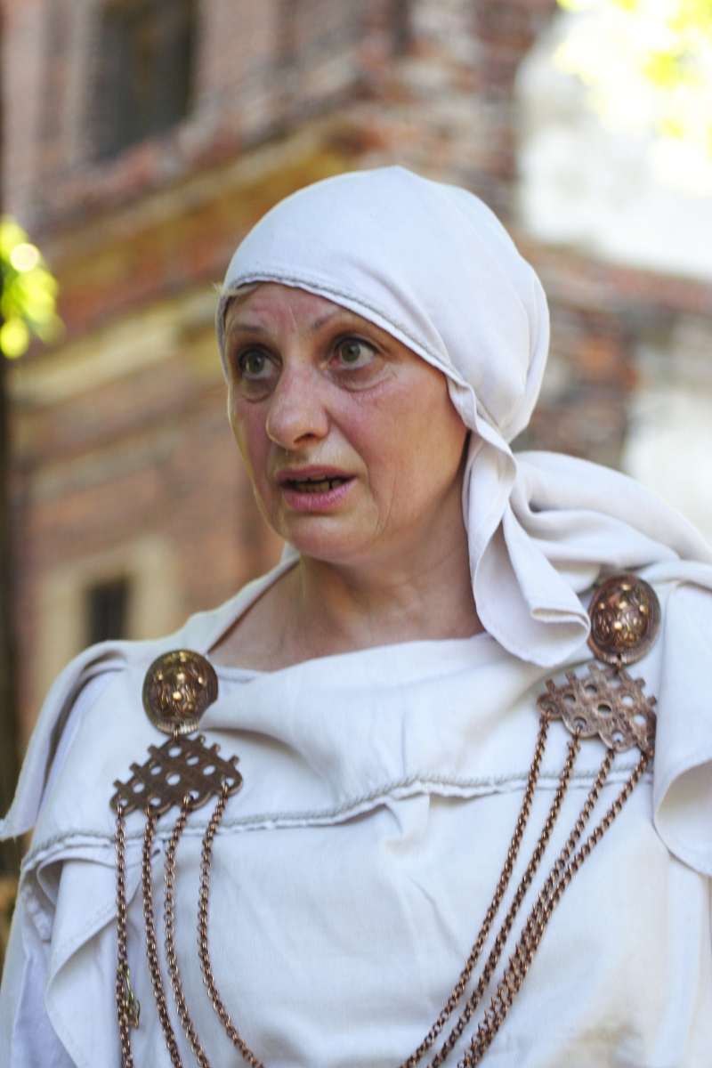 A woman wearing a white fabric headscarf and a white garment adorned with decorative bronze accents and chains stands in front of an out-of-focus background of a brick building. She appears to be speaking or reacting to something with a concerned or focused expression, reminiscent of the Dunamunde 2010 era.