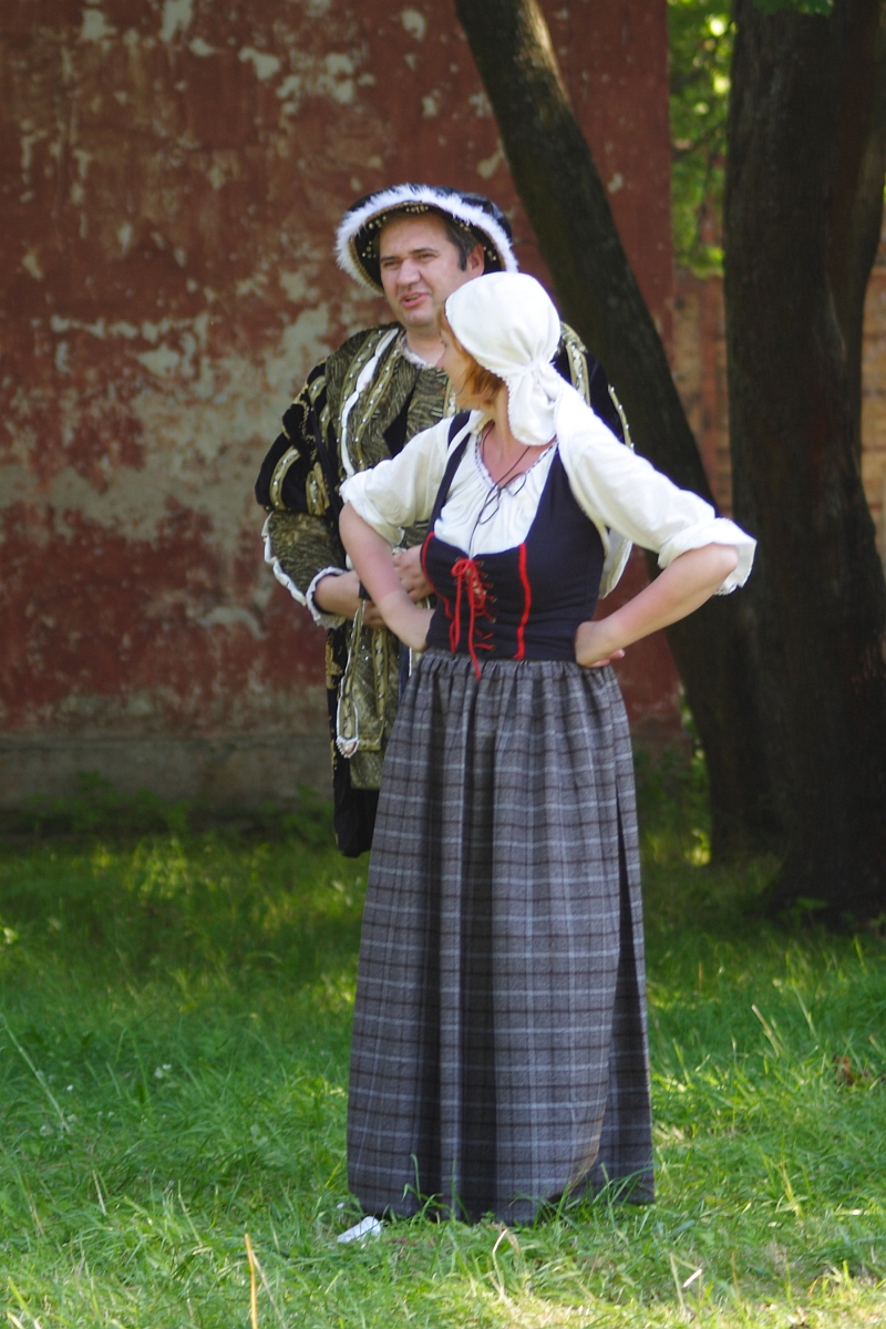 A man and woman dressed in historical costumes stand outdoors on grass, with a red and brown faded building in the background. The man, wearing an ornate outfit with a hat, and the woman, donning a long plaid skirt, white blouse, and headscarf, appear to be engaged in conversation or posing. Perhaps this scene is from the 2010 reenactment of Dunamunde.