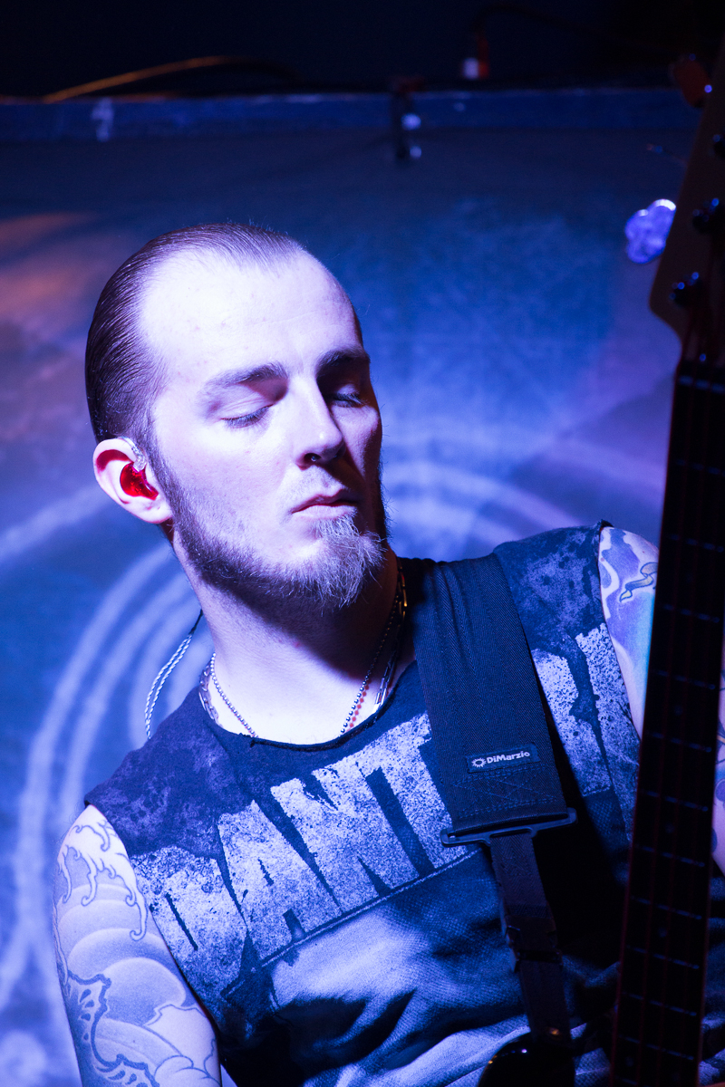 A man with a shaved head and goatee stands with eyes closed while playing an electric guitar, embodying the spirit of folk metal. His tattoos are visible on both arms as he wears a sleeveless shirt with partially visible text. The blue and purple lighting adds to the dramatic effect, reminiscent of an Eluveitie band performance.