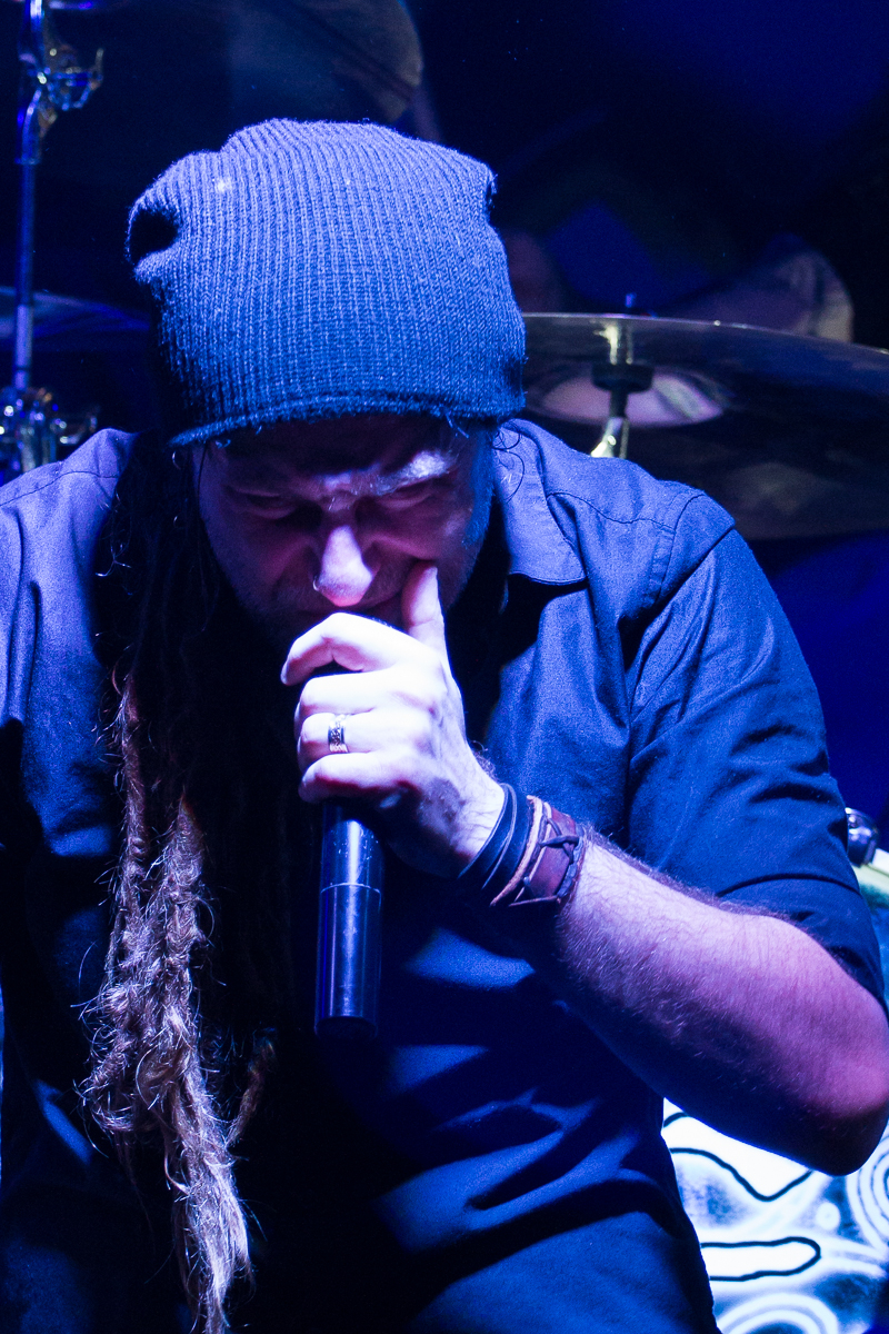 A person with long, braided hair and a beard, reminiscent of Eluveitie's style, wears a knitted beanie and dark shirt, passionately singing into a microphone. Their eyes are closed as they clench their left hand near their mouth. The background features blurry, dark lighting and parts of a drum set.