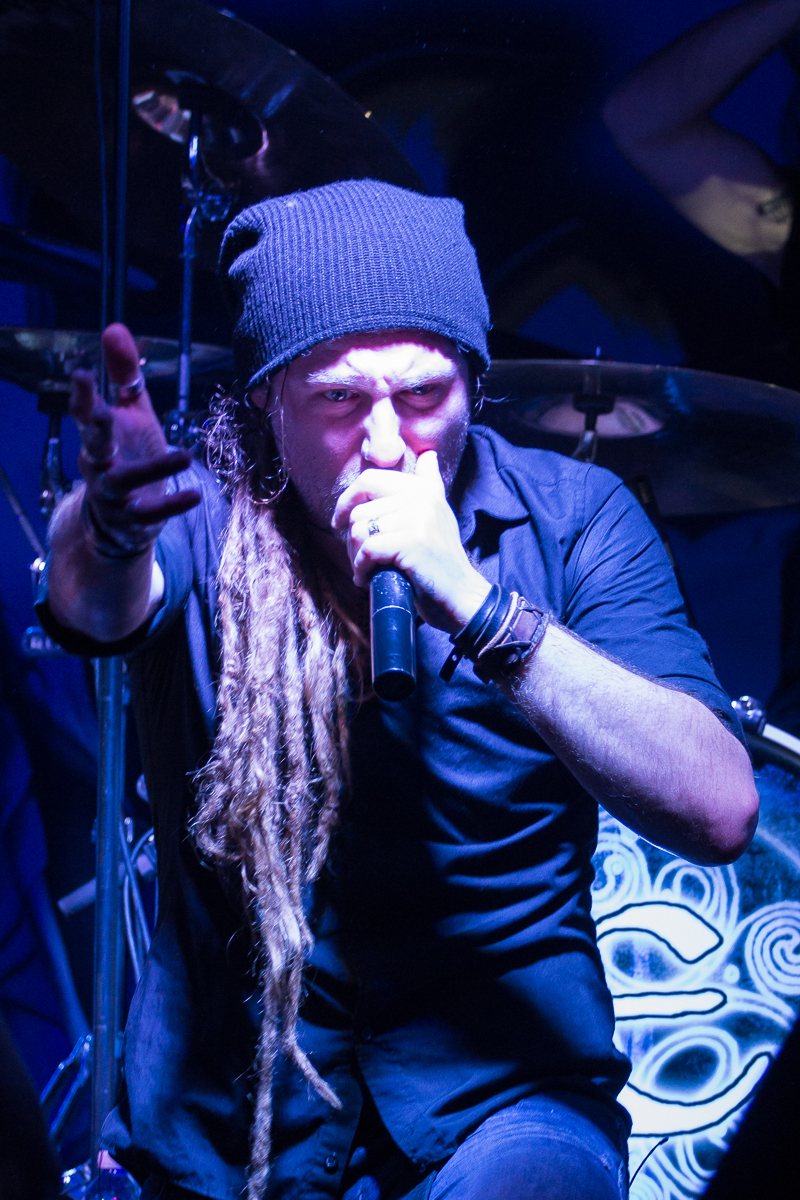 A man with long dreadlocks, wearing a black beanie and black clothing, is passionately singing into a microphone. His left arm is outstretched, pointing towards the audience. The background includes part of a drummer from Eluveitie, with drums featuring artistic designs. The lighting creates a dramatic scene.