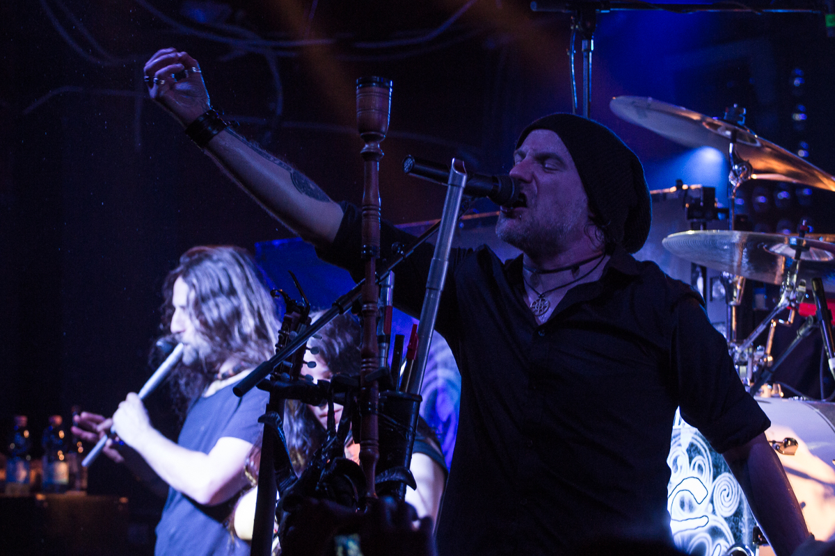 A musician in a beanie passionately sings into a microphone while playing the bagpipes on stage, reminiscent of Eluveitie's style. Another band member with long hair plays the flute in the background. The stage is dimly lit with blue and purple lights, creating a moody atmosphere. Drum set and other instruments are visible.
