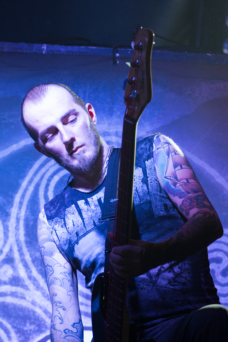 A musician with a beard and tattoos on both arms plays a bass guitar on stage with Eluveitie, a renowned Swiss folk metal band. He wears a sleeveless shirt, revealing detailed, colorful tattoos. The background is illuminated in blue and purple lights, highlighting the intricate designs on his arms. His eyes are closed, conveying deep focus.
