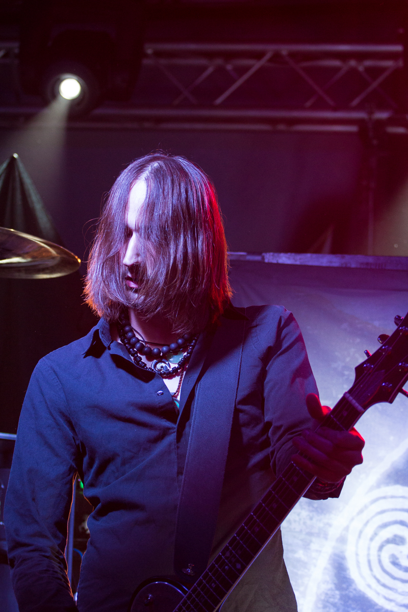 A guitarist with long, brown hair partially covering his face plays an electric guitar on stage. He's wearing a black shirt and several necklaces. Bright stage lighting highlights his figure against a backdrop displaying abstract swirling patterns, enhancing the live rock concert vibe reminiscent of Eluveitie's performances.
