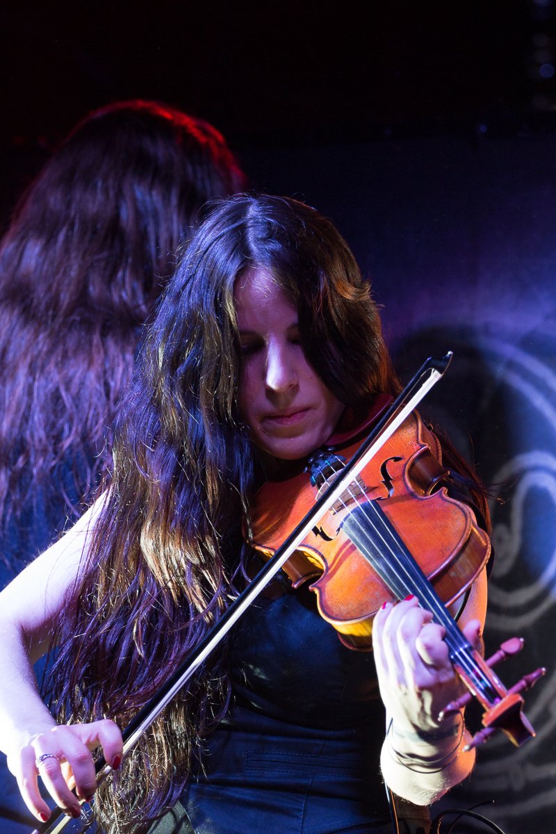 A musician with long, dark hair plays a violin on stage, focusing intently on the instrument. She wears a sleeveless black top and is bathed in colorful stage lighting, evoking the energy of a Swiss folk metal performance. Another person with long hair is partially visible in the background, facing away from the camera.