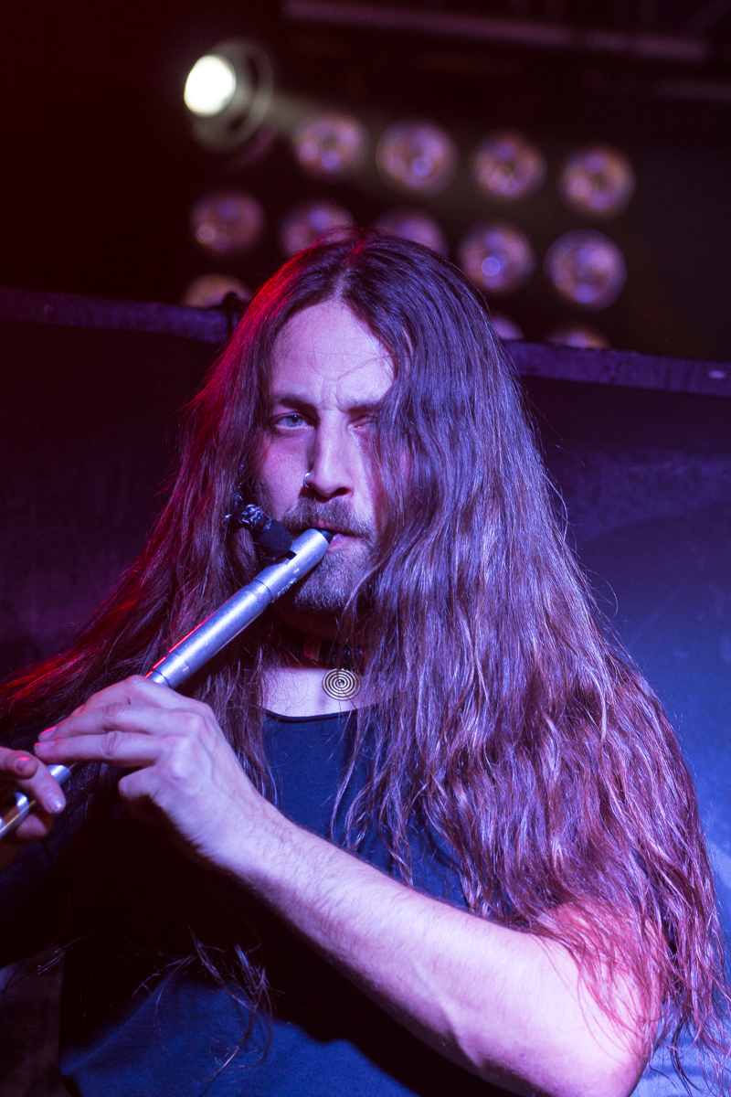 A musician with long brown hair plays a silver flute on stage, embodying the spirit of Swiss folk metal. Wearing a black shirt and illuminated by stage lights, he is intensely focused. Hints of purple and red lighting enhance the scene, where parts of the stage setup for Eluveitie are visible in the background.