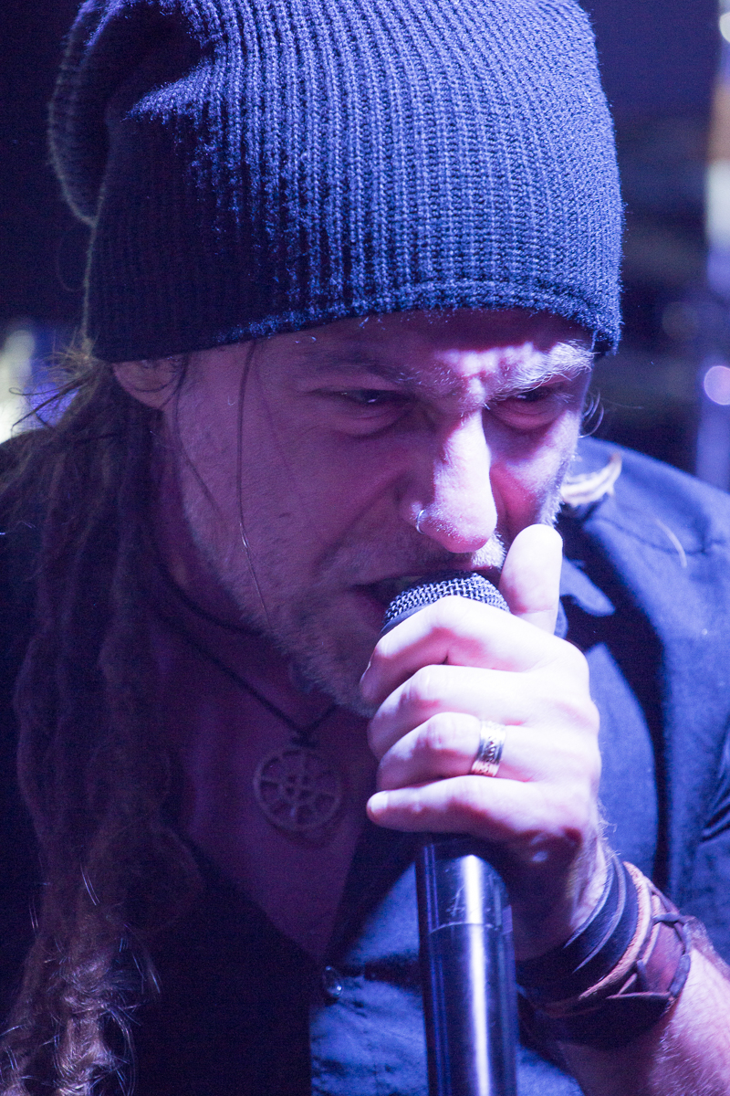A person with long, braided hair wearing a black knit beanie passionately sings into a microphone, reminiscent of Eluveitie's energetic performances. They are dressed in a dark shirt and have a ring on their finger and multiple bracelets on their wrist. Their expression is intense and focused, with the background blurred.