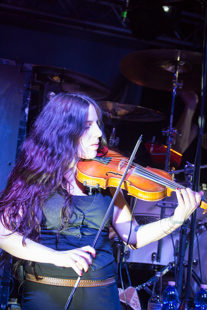 A woman with long, wavy dark hair plays a violin on stage. She is wearing a black sleeveless top and has tattoos on her left forearm. Behind her, drums and a cymbal hint that she is part of Eluveitie, a renowned Swiss folk metal band. The stage glows with blue and purple lighting.