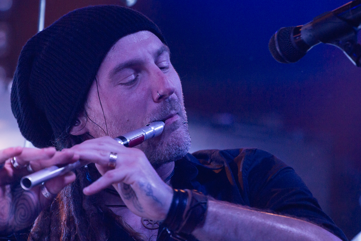 A man with long hair, wearing a black beanie and a black shirt, plays a silver flute with closed eyes. He has tattoos on his hands and wears several rings and bracelets. A microphone is positioned near him. The background is blurred with blue and purple lighting, suggesting a folk metal stage performance reminiscent of Eluveitie.