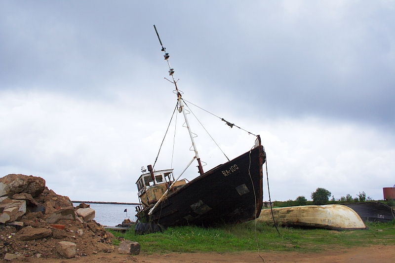 A weathered, old fishing boat is stranded on the shore in Engure, tilted slightly to one side. The sky overhead is overcast with dark, gray clouds. Piles of rocks and dirt surround the boat, emphasizing its abandonment. A smaller, overturned vessel lies nearby, adding to the scene of neglect.