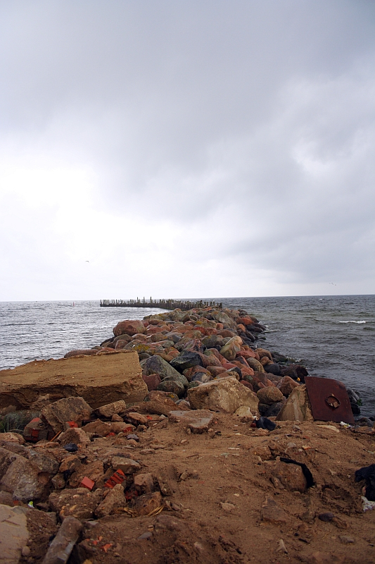 A rugged, rocky pier in Engure juts into a tumultuous sea under a gloomy sky, with colorful boulders forming a barrier against the waves.