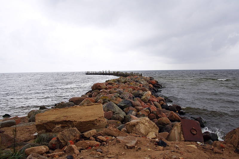 A rocky breakwater extends into the sea at Engure, comprised of large, multicolored stones. The water is calm, with gentle waves lapping against the rocks. The sky is overcast with thick, gray clouds. In the foreground, additional stones and some debris rest on sandy ground.