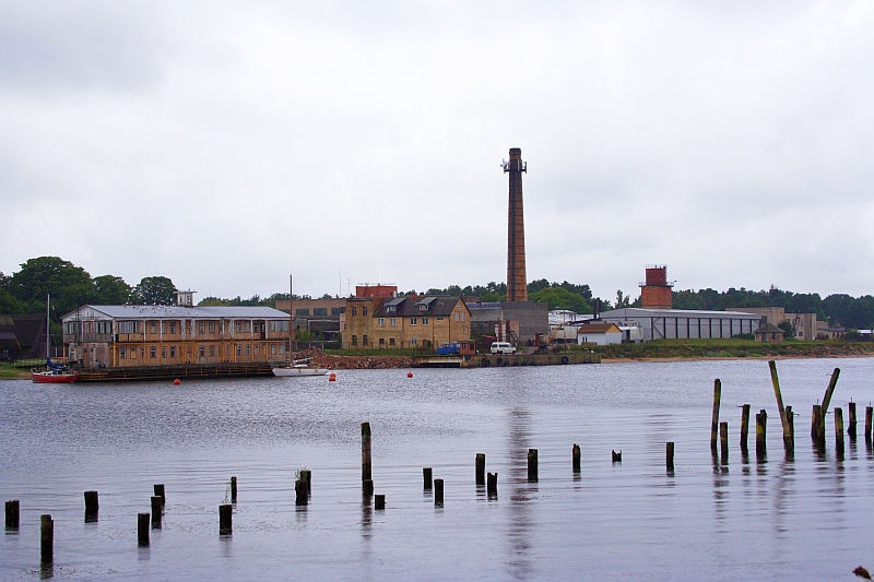 A tranquil overcast day by the riverside in Engure, featuring an industrial landscape with a prominent chimney, older buildings, and mooring posts in the foreground, reflective of a bygone era's