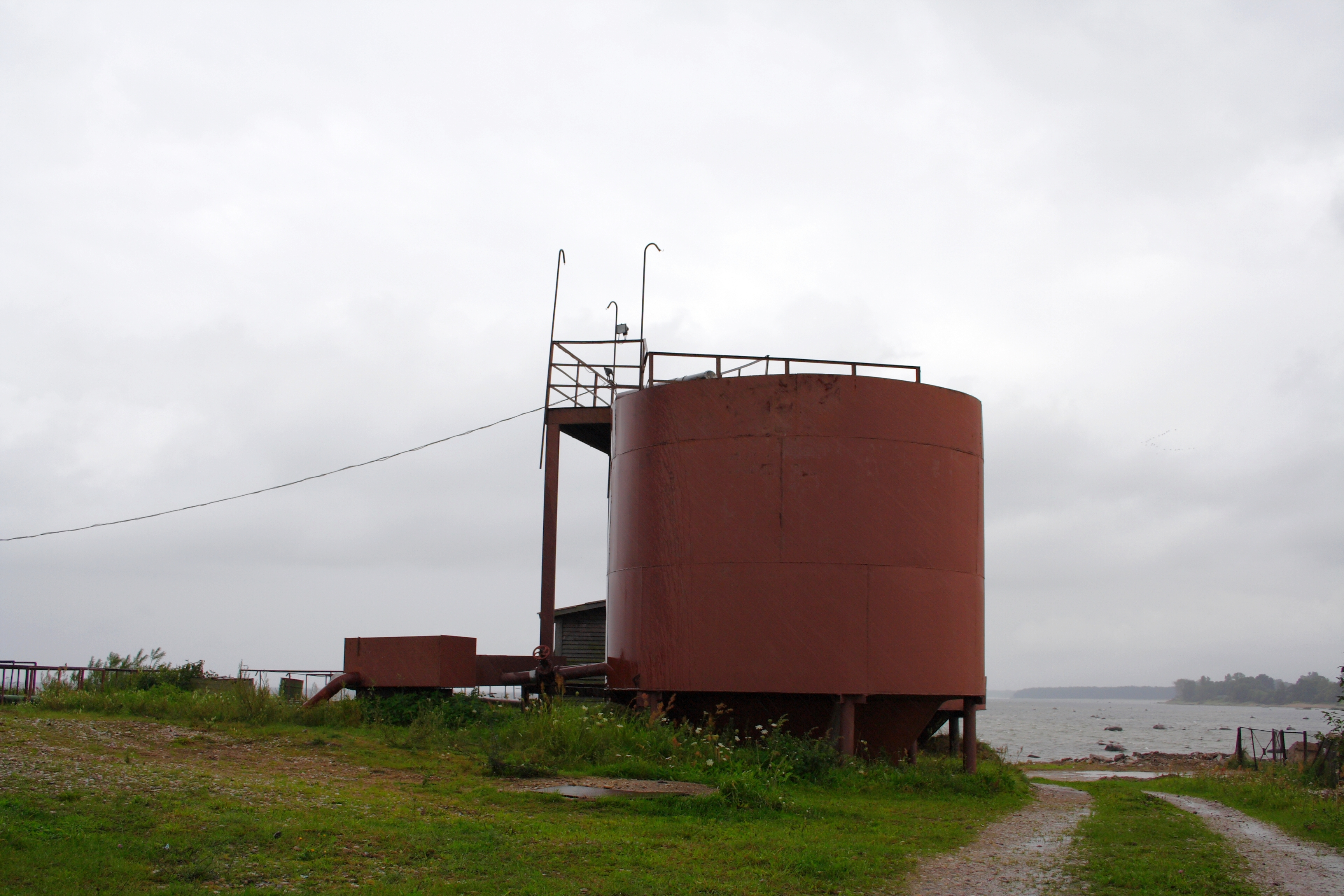 A large, rusty cylindrical tank with a metal staircase stands under an overcast sky near the Engure body of water, conveying a sense of industrial abandonment and solitude.