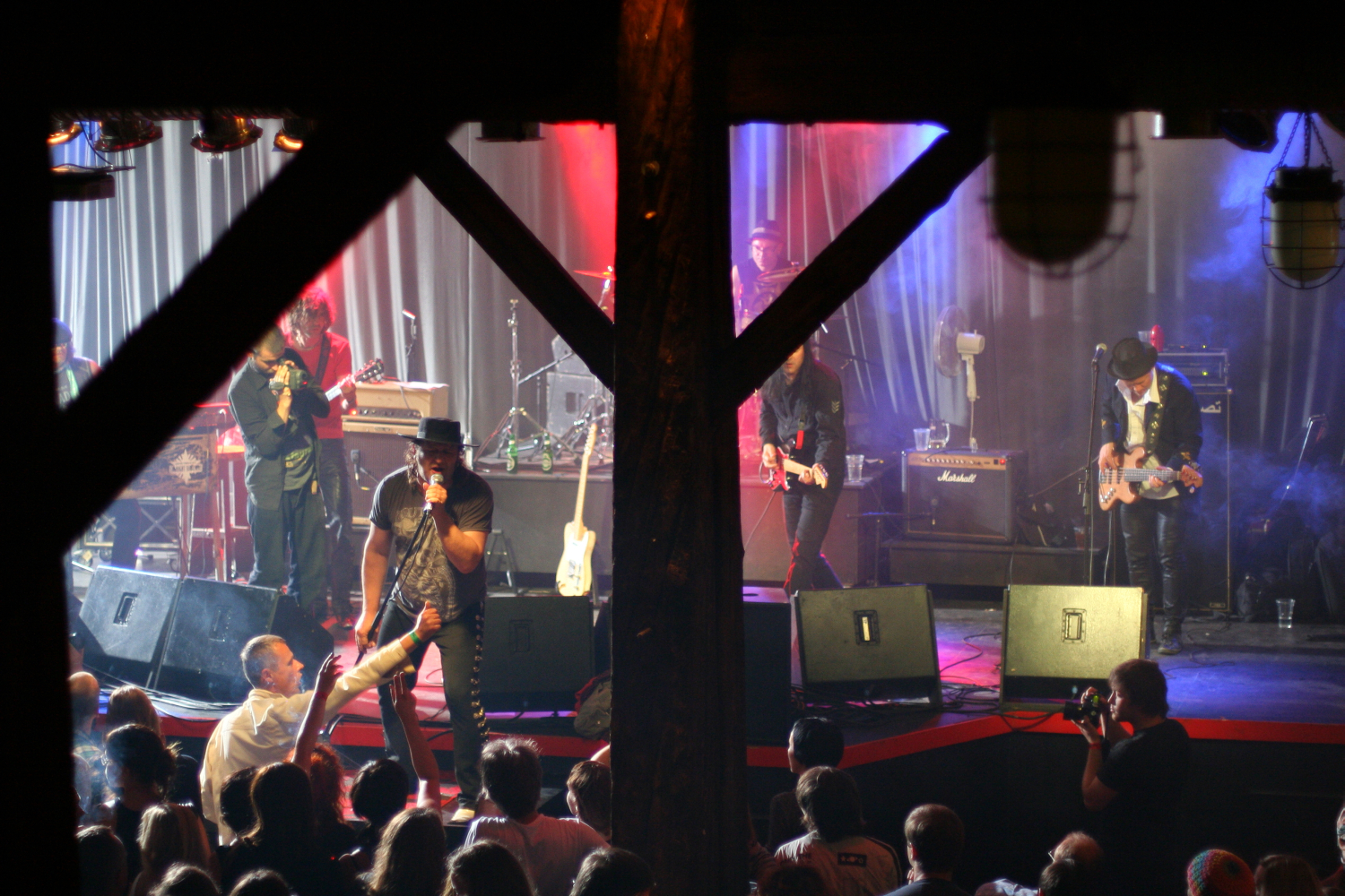 At the 2011 Fontaine Festival, a rock band performs on stage with vibrant red and blue lighting. The lead singer, wearing a black hat, engages with the audience while the guitarist and bassist play alongside. A wooden beam frames the stage. An audience watches the performance, and a photographer captures the moment.