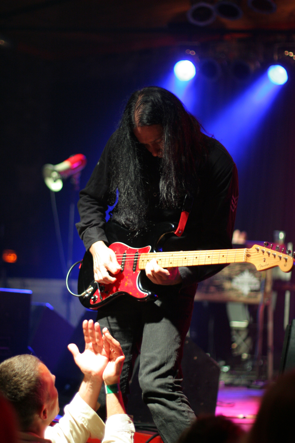 A guitarist with long black hair, dressed in black, is passionately playing an electric guitar on stage during the 2011 Fontaine Festival. He is leaning forward towards the audience, who are reaching out towards him. The stage is dimly lit with blue and purple lights, creating a vibrant atmosphere.