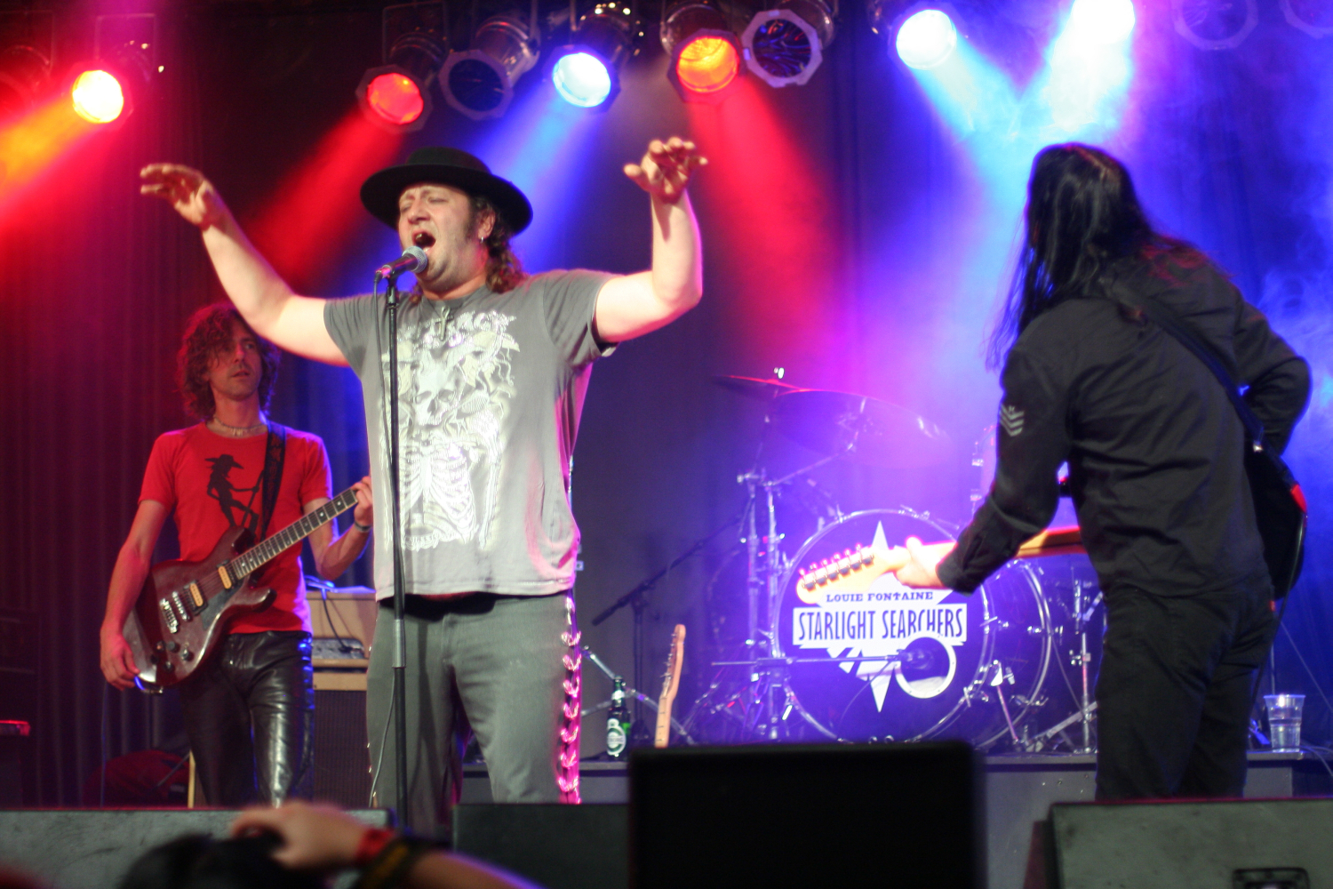 At the 2011 Fontaine Festival, a man in a hat and graphic t-shirt sings passionately into a microphone on stage, with arms raised. Another musician to the left plays an electric guitar, and a third man to the right readies another instrument. The backdrop features colorful lights and a drum set with "Starlight Searchers" written on it.