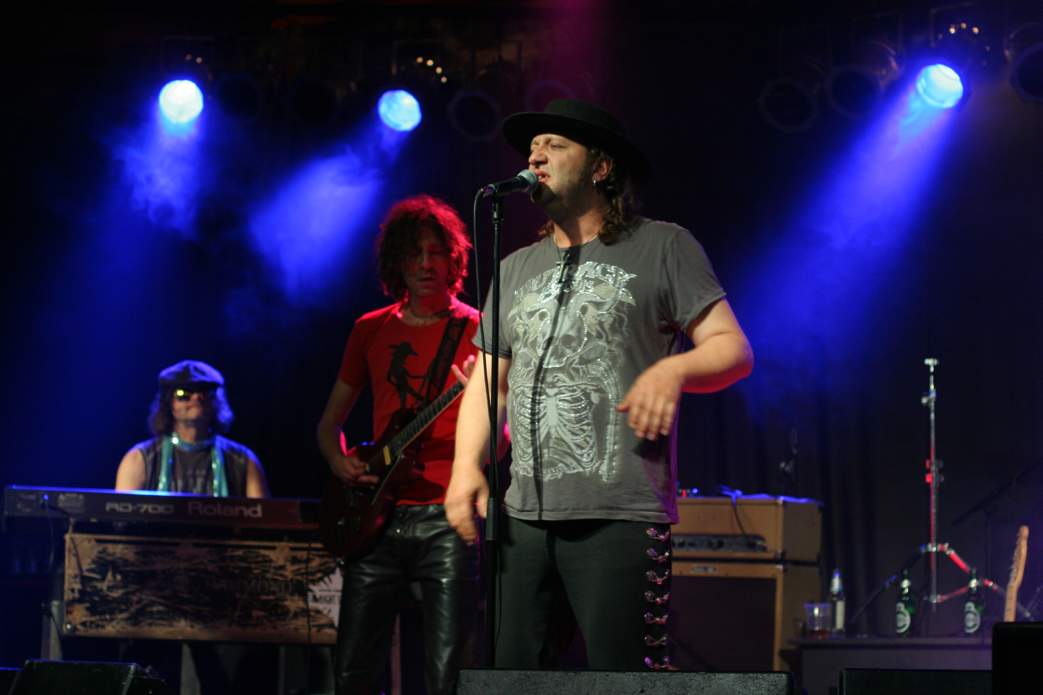 At the 2011 Fontaine Festival, a rock band performs on stage under blue and purple lighting. The lead singer, wearing a black hat and gray t-shirt, sings into a microphone. To his left, a guitarist with curly hair and a red shirt plays. A keyboardist and drum set are visible in the background.