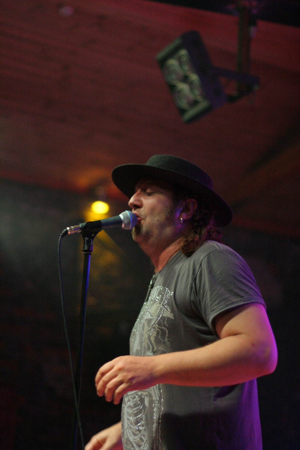 A person with long hair wearing a black hat and a gray T-shirt sings into a microphone on a dark stage at the Fontaine 2011 festival. They are bathed in a mix of stage lights, including a focused yellow light above their head. The background is dimly lit, with a visible light rig hanging from the wooden ceiling.
