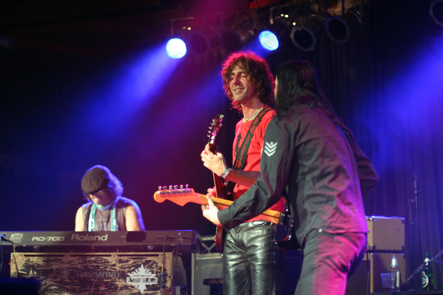Three musicians perform on stage at the Fontaine festival under colorful lighting. One plays a keyboard on the left, wearing a hat. The second musician, in the middle, plays an electric guitar and is smiling. The third musician, on the right, faces away from the camera, playing another electric guitar.