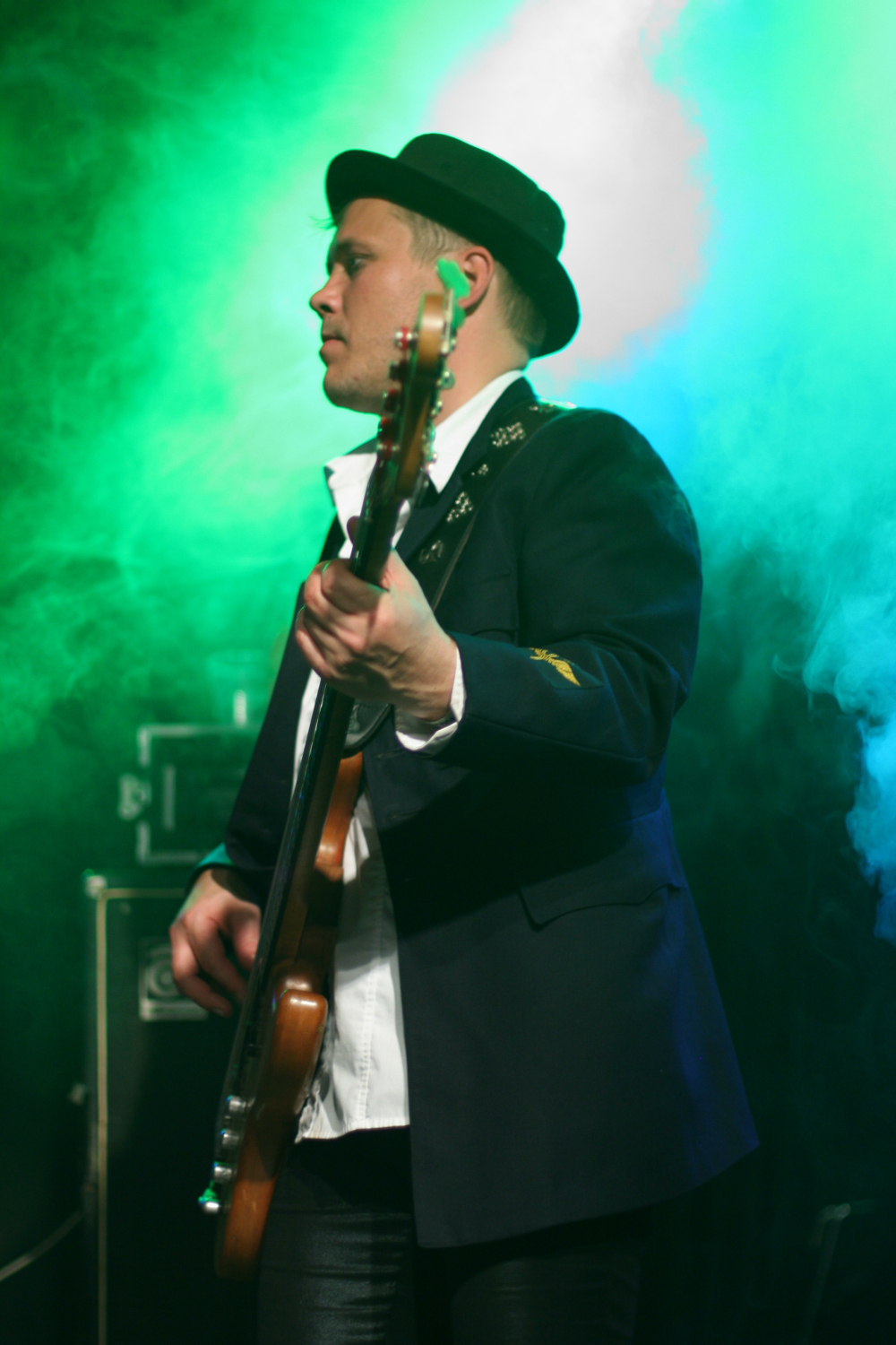 A musician in a black fedora and dark blazer plays a guitar on stage at the 2011 Fontaine festival. He stands under green and blue lights, partially obscured by stage smoke. The musician looks focused, with his gaze directed to the side. The background shows stage equipment in soft focus.