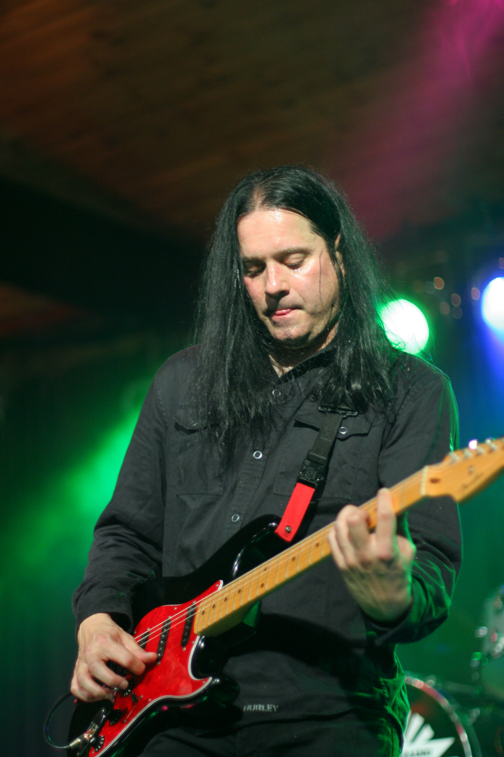 At the 2011 Fontaine festival, a musician with long black hair plays an electric guitar on stage. He is wearing a black shirt and is focused on his performance. The guitar has a red strap, and green and blue stage lights in the background create a vibrant atmosphere.