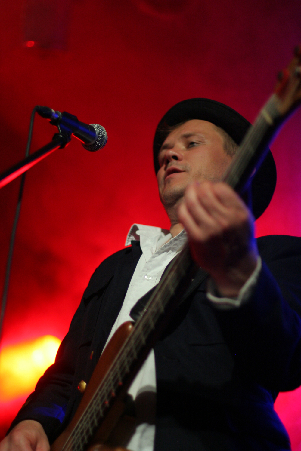 A musician wearing a black hat and a black jacket over a white shirt plays an electric guitar on stage at the 2011 festival. The background is illuminated with vibrant red and yellow lights, and a microphone is visible in front of the performer. The atmosphere suggests an energetic live music performance.