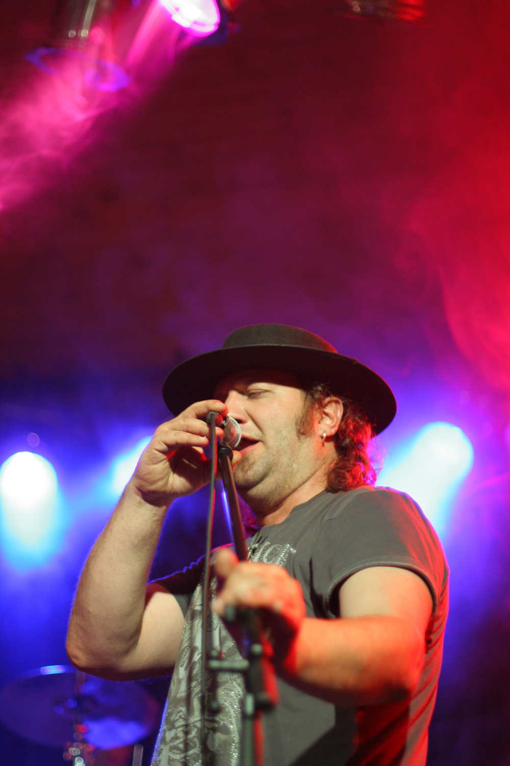 At the Fontaine Festival 2011, a person with a beard wearing a black hat and a gray t-shirt is singing into a microphone on stage, illuminated by vibrant red and blue stage lights. Their eyes are closed as they hold the microphone stand with their right hand, conveying emotion in their performance.