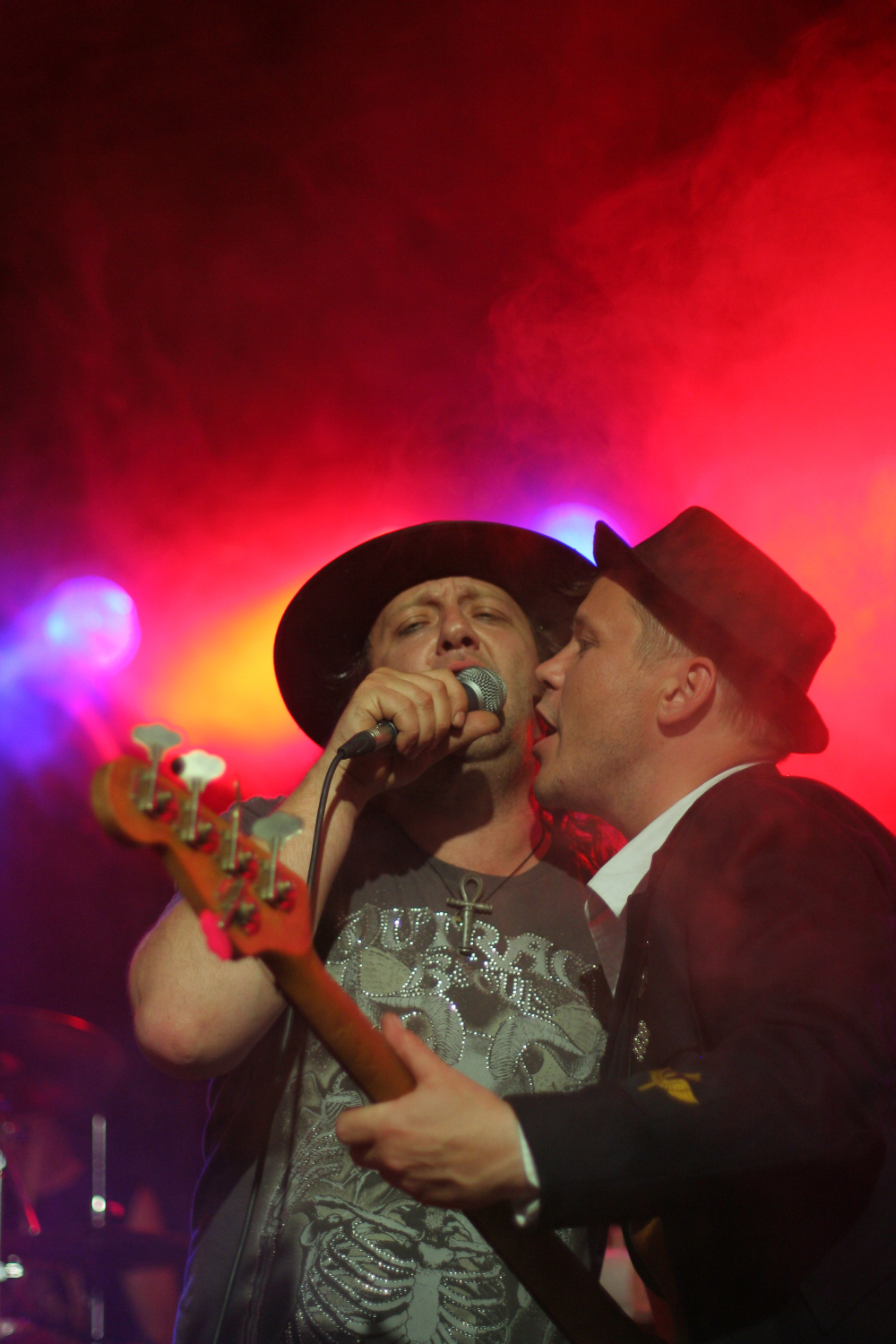 Two male musicians perform on stage amid vibrant, colorful lights at the Fontaine Festival 2011. One, wearing a black hat and graphic t-shirt, sings into a microphone. The other, in a suit and dark hat, leans in close, holding a guitar and also singing. The background is illuminated with orange, red, and purple lighting.