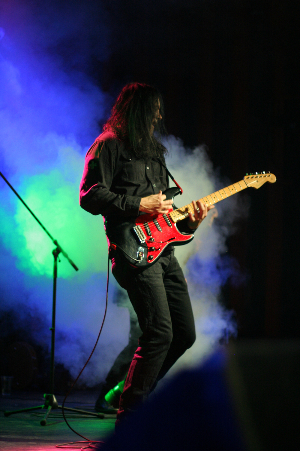 A guitarist with long hair performs energetically on stage at the Fontaine Festival 2011, playing a red electric guitar. He wears a black shirt and pants. Colorful stage lights in green and blue, along with thick white smoke, create a dramatic background. A microphone stand is visible to the left.