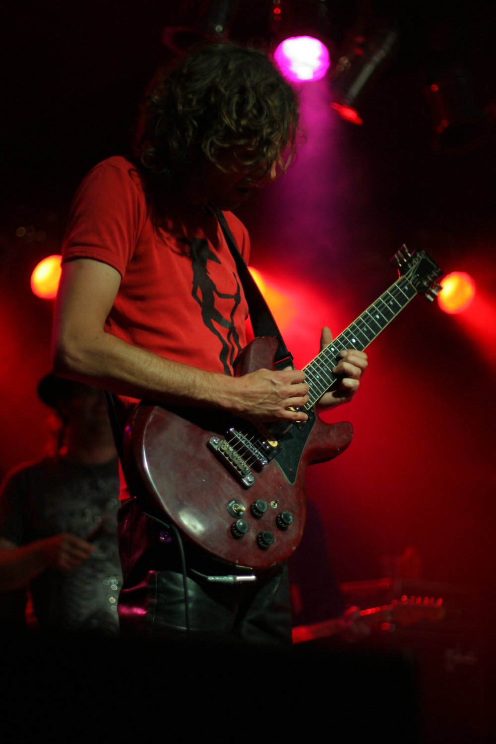 A guitarist with long curly hair, wearing a red shirt and black pants, plays an electric guitar under red and purple stage lights at the Fontaine Festival 2011. Another person stands in the background, partially visible. The stage is dimly lit with a blurred audience in the background.