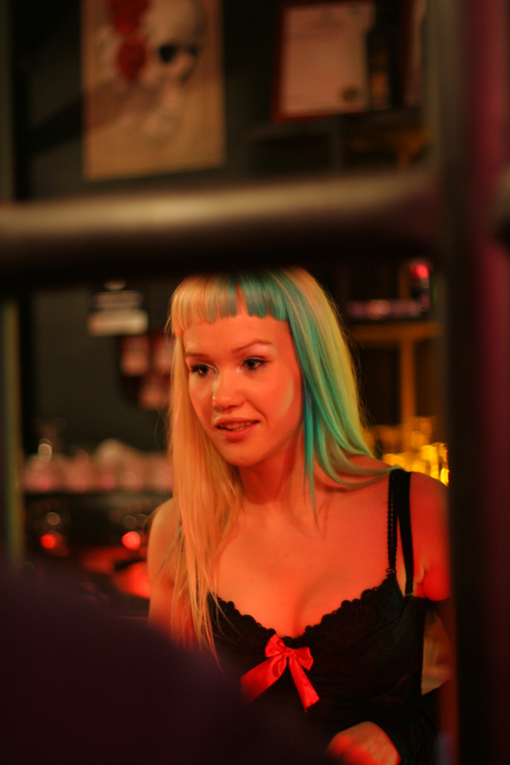 A woman with two-toned straight hair, blonde and green, is photographed indoors under warm, dim lighting. She has blunt bangs and is wearing a black top with a pink bow. In the slightly blurred background featuring decor elements and various bar items, a poster for the Fontaine festival 2011 can be seen.