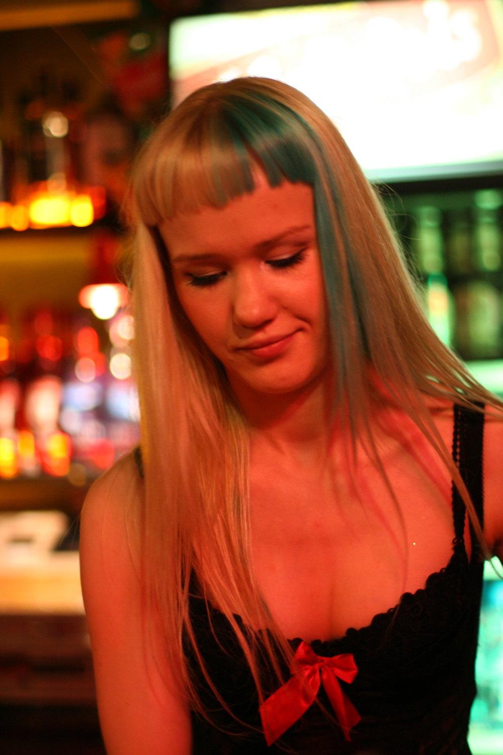A person with long blonde hair featuring blue highlights and blunt-cut bangs is looking down and slightly smiling. They are wearing a black top with a red ribbon. The background is illuminated with warm, colorful lights, suggesting a lively indoor setting like the 2011 Fontaine Festival or a bar.