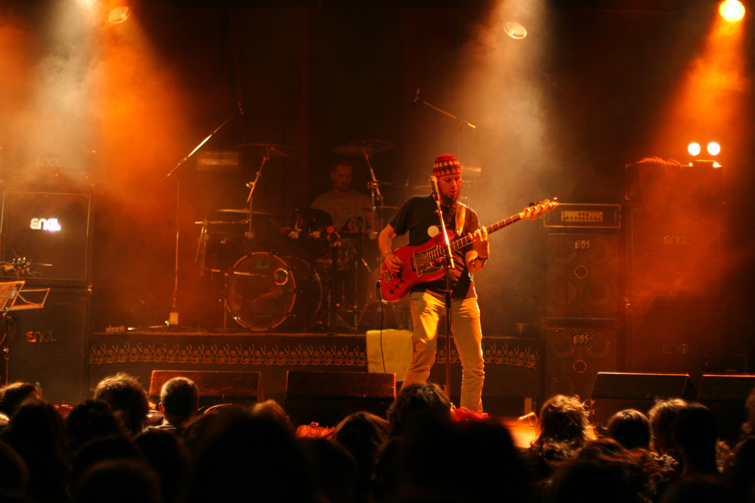 A guitarist performs on stage under warm amber lighting during the Fontaine Festival 2011, wearing a hat and casual clothing. A drummer is visible in the background, playing amidst smoke effects. Amplifiers and equipment are positioned around the stage. An audience watches from the foreground, silhouetted by the stage lights.