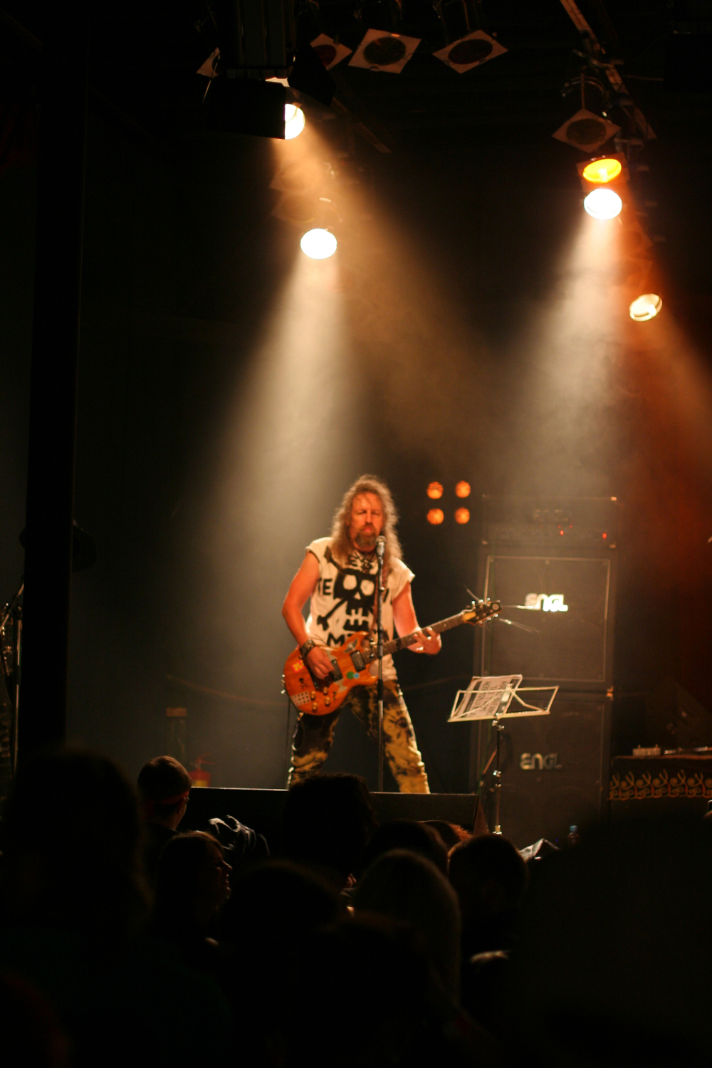 A guitarist with long hair is performing on stage under warm stage lights at the 2011 Fontaine Festival. He is wearing a white sleeveless shirt and camouflage pants, playing an electric guitar. Behind him, amplifiers and a music stand are visible. The audience is partially visible in the foreground, watching the performance.