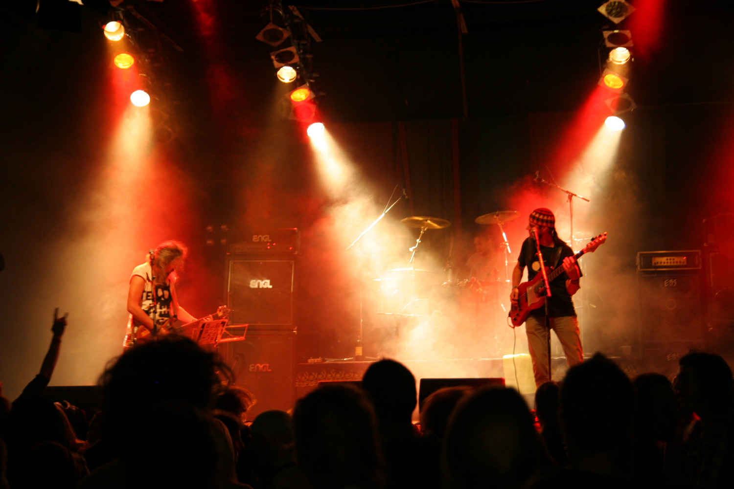 At the 2011 festival, a rock band is performing on stage with dramatic red and yellow lighting illuminating the scene. The guitarist on the left is playing and standing near a microphone, while the bassist on the right wears a headband. A drum set is visible in the background. A crowd watches from the foreground.