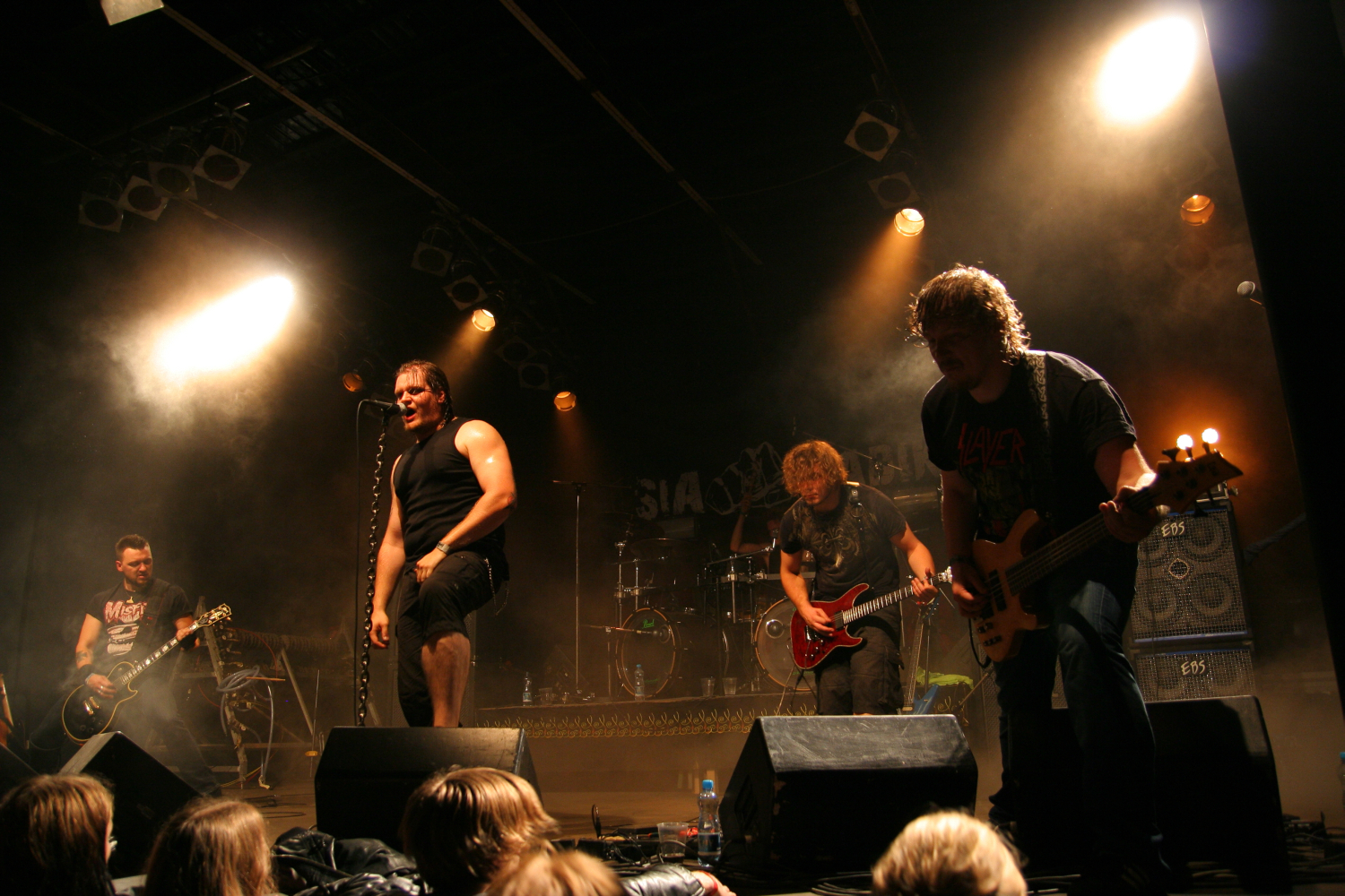 At the Fontaine festival 2011, a band performs energetically on stage with bright spotlights and fog adding to the ambiance. The lead singer, wearing a black tank top, leans into the microphone. Two guitarists and a bassist, all in casual clothes, play passionately, while the drummer is partially visible in the background.