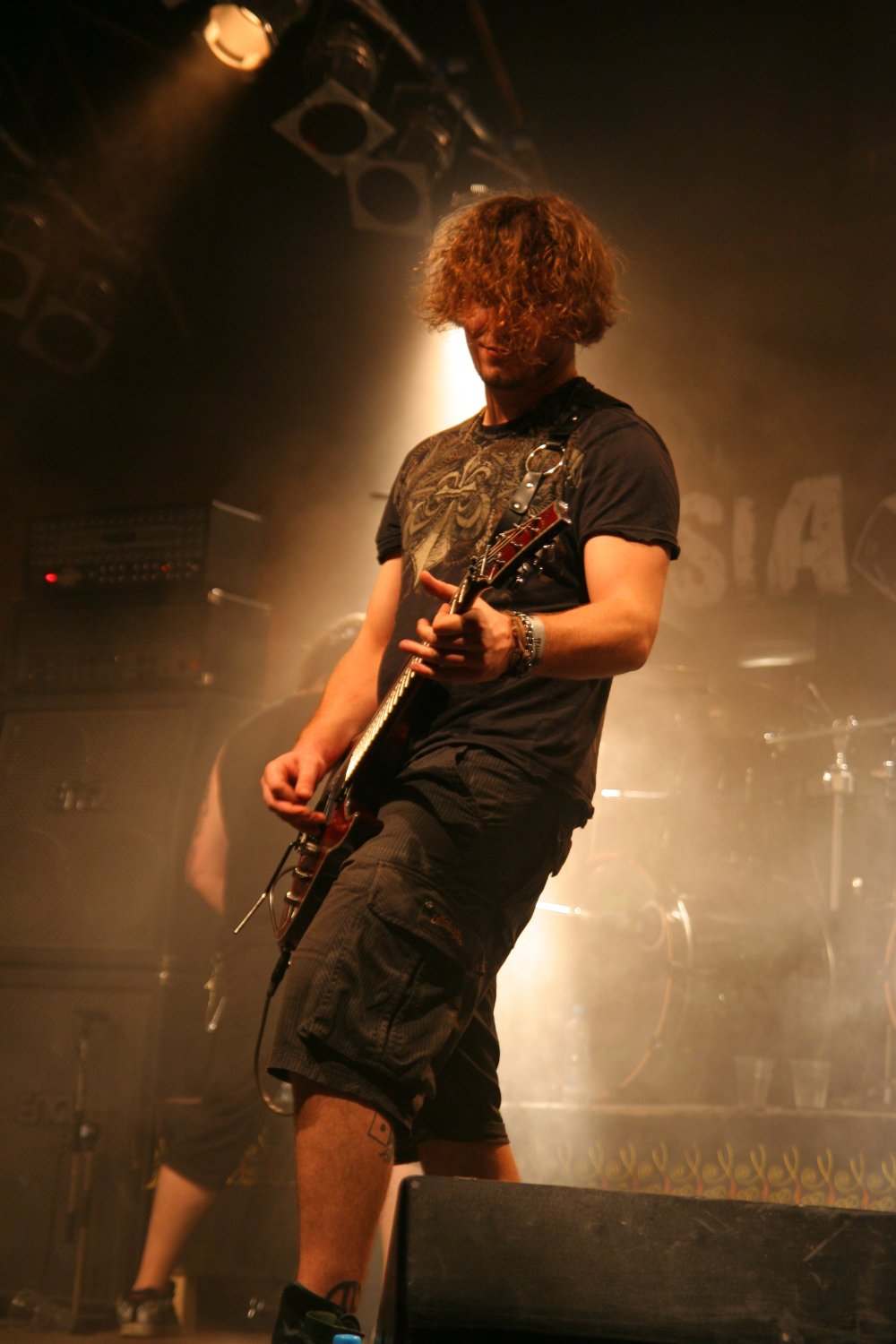 A musician with curly hair, wearing a dark t-shirt and shorts, energetically plays an electric guitar on stage at the 2011 Fontaine Festival. The stage is dimly lit with spotlights and smoke effects. Amplifiers and drum equipment are visible in the background, along with indistinct band signage on the back wall.