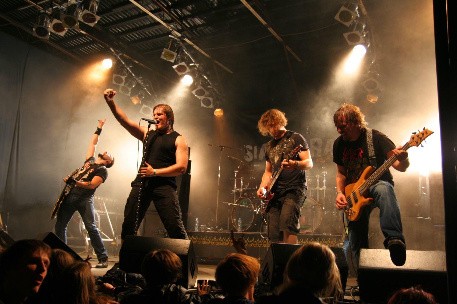 A rock band performs energetically on stage at the Fontaine festival 2011. The lead singer holds a microphone and belts out a song while raising a fist. Other band members play electric guitars and a bass, and one raises an arm in the air. Stage lights shine from above, creating a dramatic atmosphere as the crowd watches.