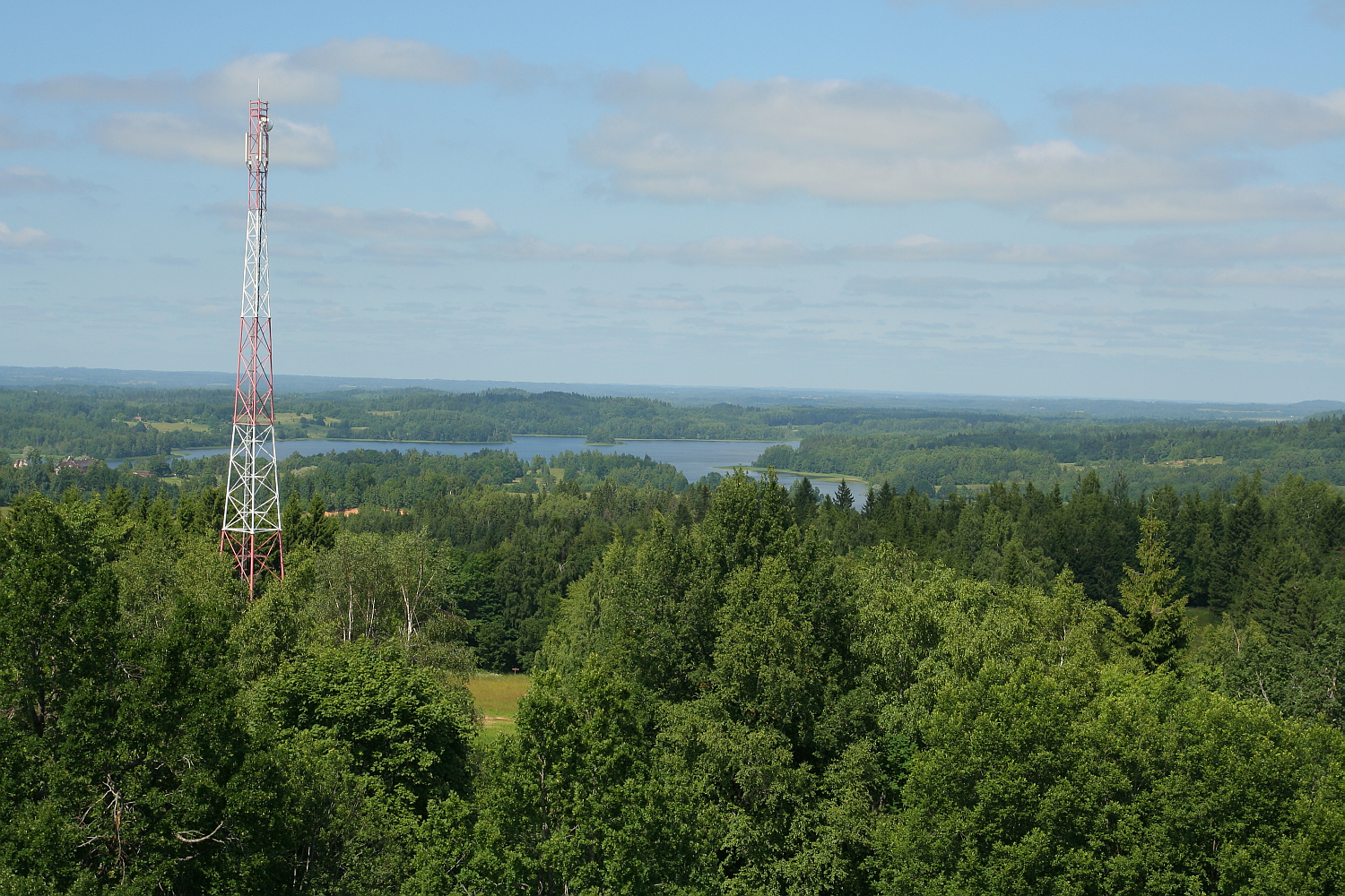 A towering red and white communications mast rises above a lush green forest near Gaiziņš with a serene lake visible in the distance under a clear blue sky.