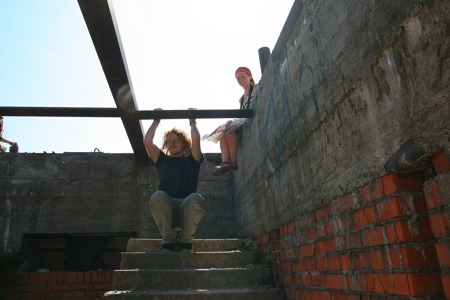 A man is crouched on a set of concrete stairs beneath a steel beam in a partly constructed building, holding onto a horizontal beam above him. A woman sits at the edge of a concrete wall beside him. The walls have visible graffiti, and the sunlight contrasts with the shadows inside, like something out of Gaiziņš' realm.